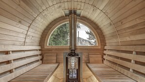 A sauna room with a wooden interior, a central sauna unit, and a large arched window providing a view of trees and mountains.