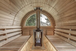 Ein Sauna-Raum mit Holzvertäfelung, einer zentralen Saunaanlage und einem großen Bogenfenster mit Blick auf Bäume und Berge.