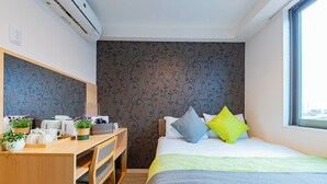 A hotel room with a bed, desk, and chairs. There is a window with a view, a wall with a floral pattern, and an air conditioning unit.