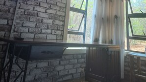 A rustic room with a brick wall, a flat-screen TV mounted above it, and a window with a view of greenery outside. -  iroCK Lodge (Victoria Falls)