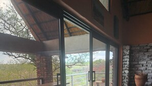 A spacious balcony with wooden furniture, a brick wall, and a large window offering a view of the landscape. -  iroCK Lodge (Victoria Falls)