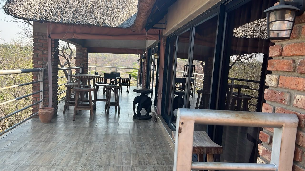 An outdoor seating area with tables and chairs, a thatched roof, and a view of the landscape.