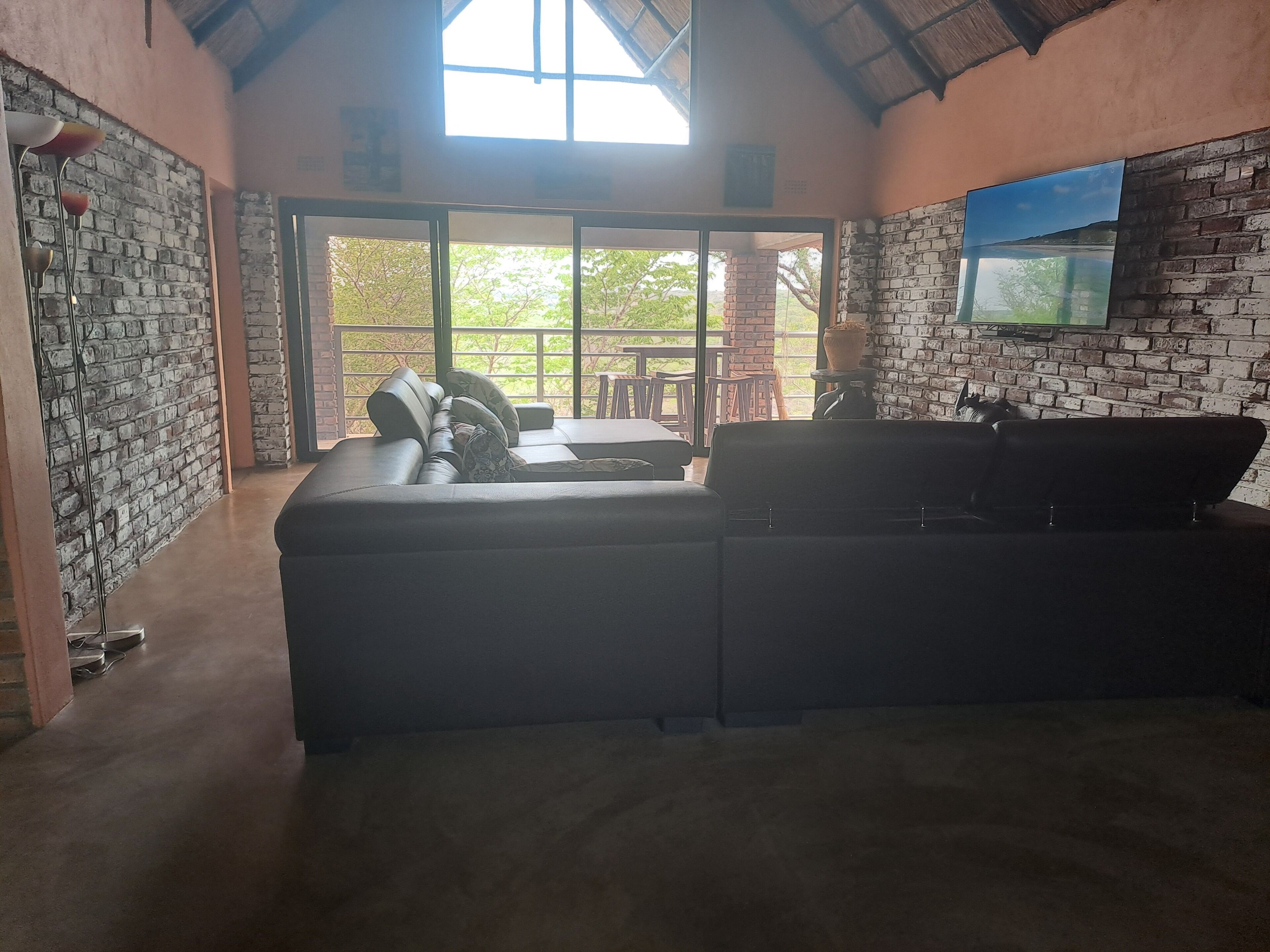 Un vaste espace de vie comprenant un grand canapé, un téléviseur fixé au mur en briques et une vue sur une terrasse extérieure, accessible par une porte vitrée.
