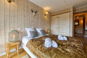 Un dormitorio con pared de madera, una cama con ropa blanca, dos almohadas, un plaid de piel sintética y un par de toallas.