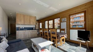 A modern living room with a sofa, a coffee table, and a TV. There is a dining area with chairs and a table. The room has wooden walls and large windows. - Résidence la Ménandière W1 (Alpe d'Huez)