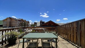Eine Holzterrasse mit Tisch und Stühlen, umgeben von einem Holzzaun, mit Blick auf eine Berglandschaft und Gebäude im Hintergrund.