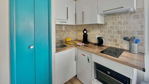 A compact kitchen with white cabinets, a teal door, a coffee maker, a kettle, and a dish rack. - Le petit Courseulles - between port and sea (Courseulles-sur-Mer)