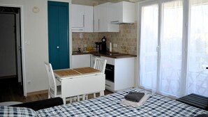 A compact kitchen with white cabinets, a blue door, and a dining area with a checkered tablecloth. - Le petit Courseulles - between port and sea (Courseulles-sur-Mer)