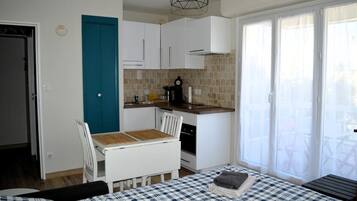 Une cuisine de taille réduite avec des meubles blancs, une porte bleue et un coin repas agrémenté d’une nappe à carreaux.