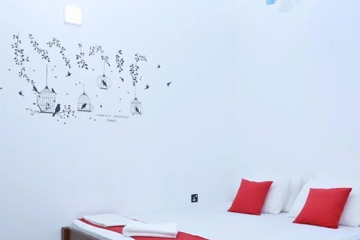 A bedroom with a wooden bed, white bedding, and red pillows. There is a wall decal of birds and a light switch.