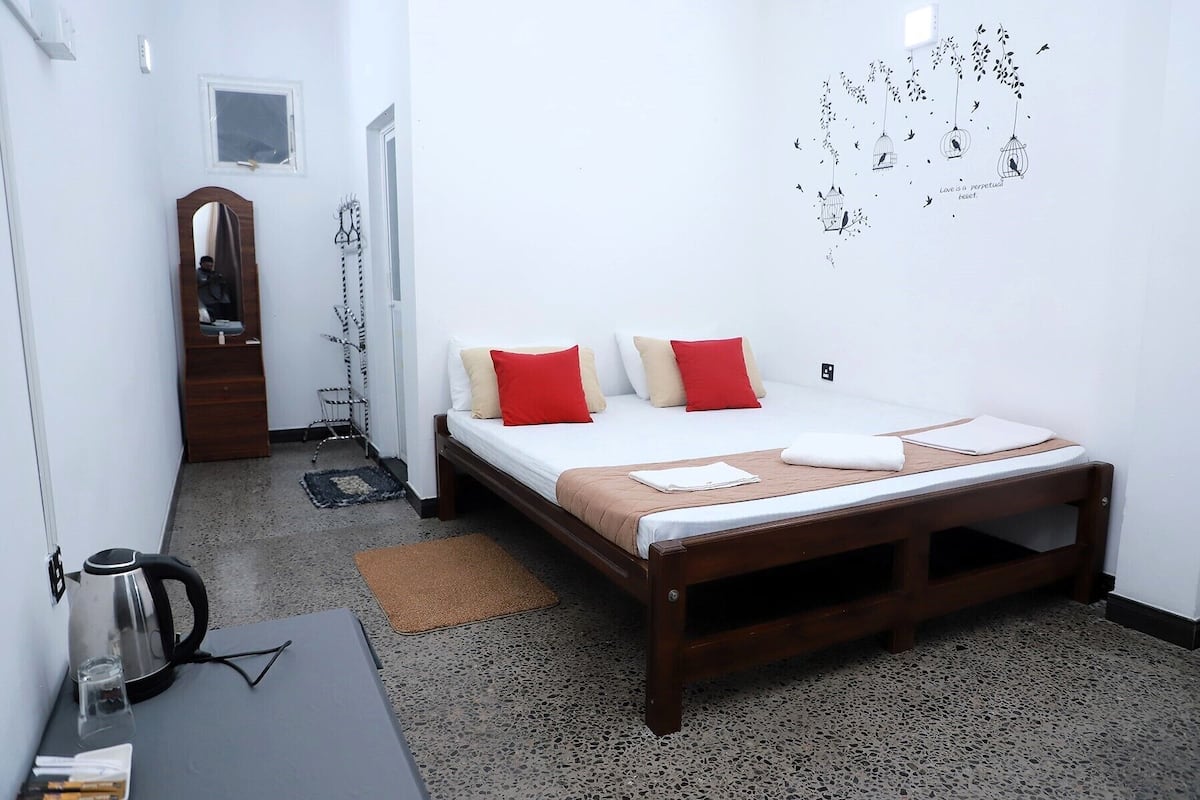 A bedroom with a wooden bed, white bedding, red and beige pillows, a small rug, a kettle, and a wall art of birds and leaves.