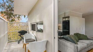 Un balcon avec des fauteuils en osier, un barbecue et une vue sur les arbres. À l’intérieur, on trouve un canapé gris et une cuisine équipée d’armoires et d’appareils électroménagers.