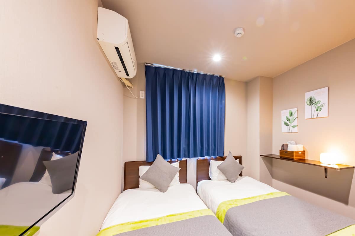 A hotel room with two beds, a bedside table, a nightstand, a wall-mounted air conditioner, a ceiling light, a window with blue curtains, and two wall-mounted plant decorations.