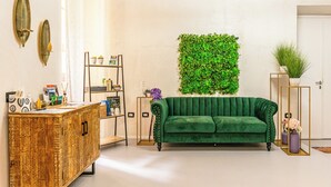 Una sala de estar con un sofá verde, un mueble de madera, una pared verde con una planta y una mesita con una planta en maceta.