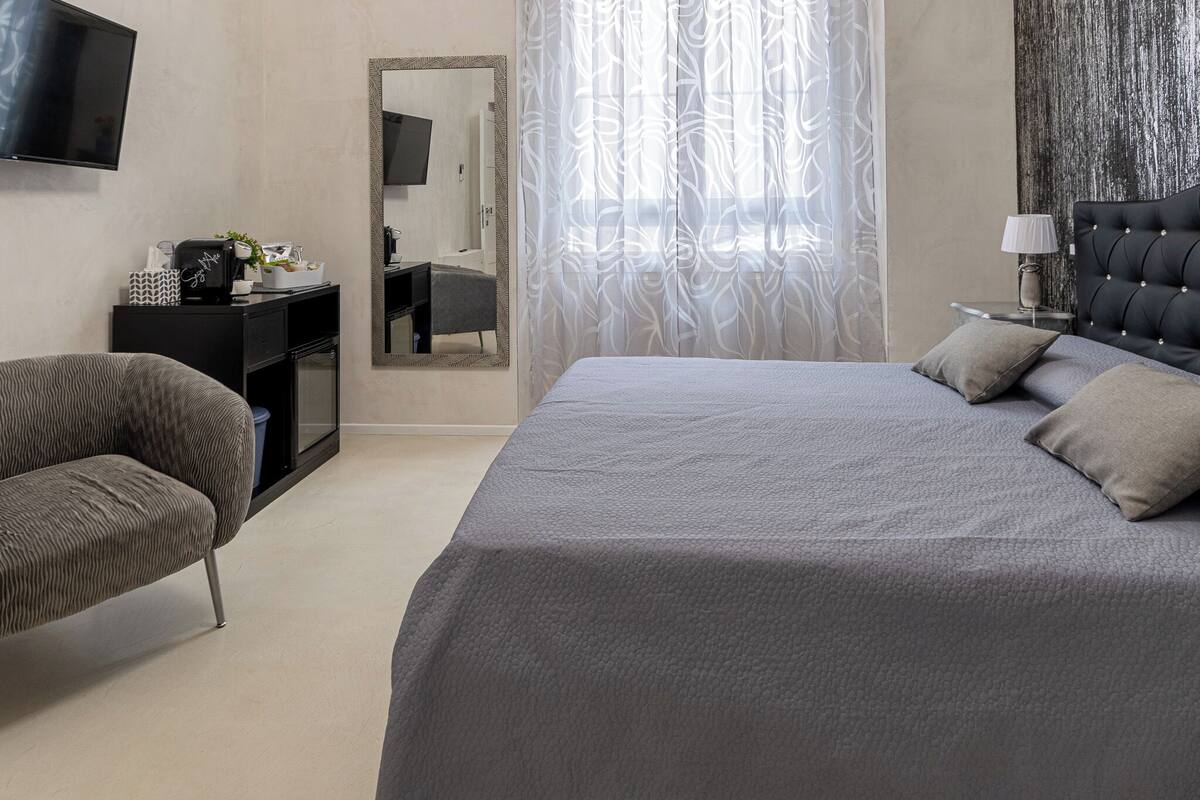 A modern hotel room with a large bed, a sofa, a desk with a mirror, and a television on the wall.