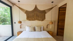 A bedroom with a large bed, two bedside tables, a hanging macrame piece, and a view of greenery outside. - Luxury 2BR Villa with Private Pool, Spacious Living & Modern Amenities (Tulum)