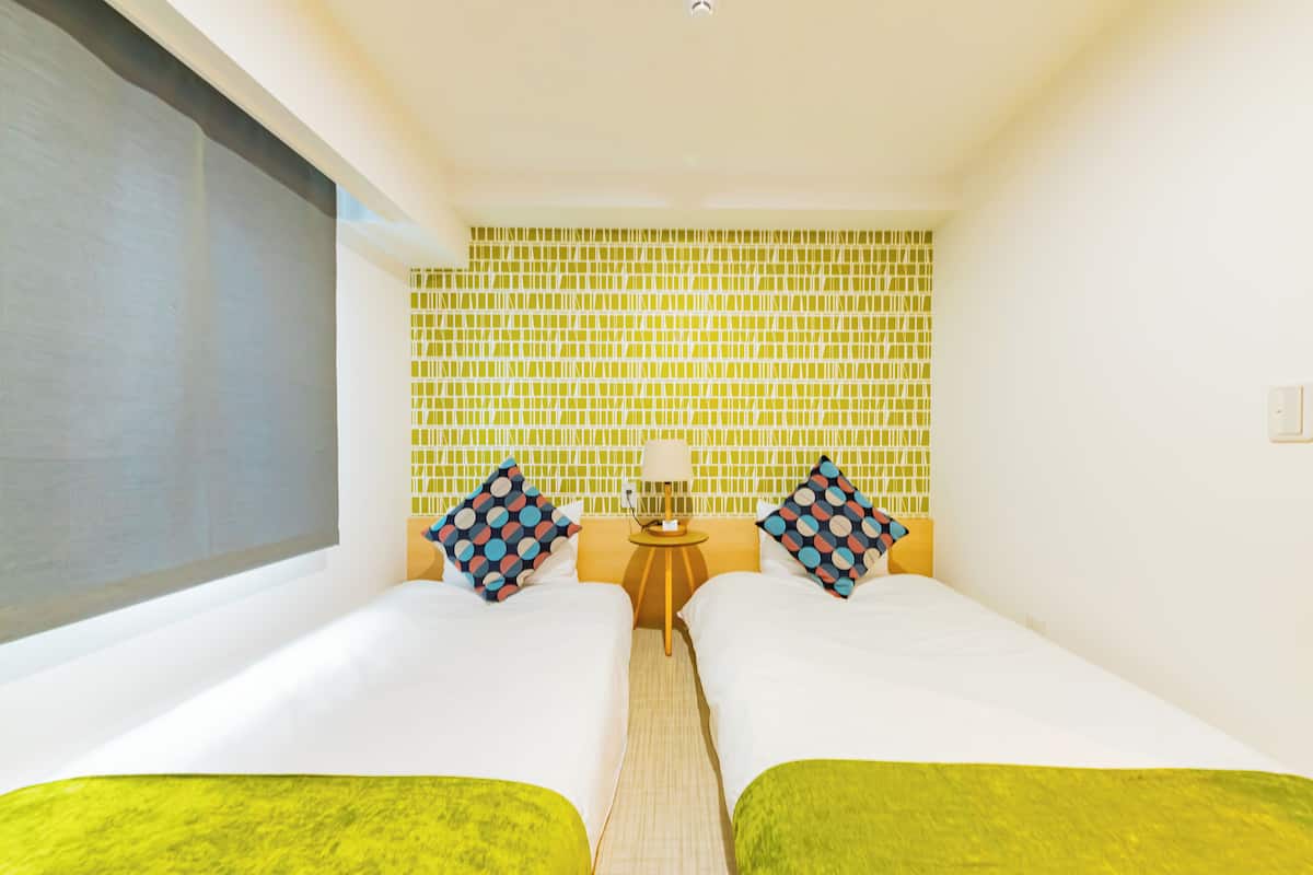 A room with two beds, a small table with a lamp, and a window.