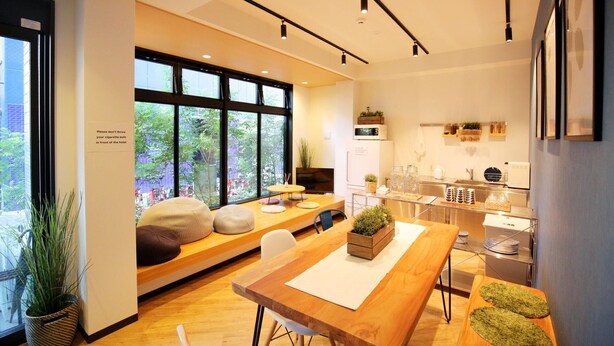 Una habitación de hotel moderna con un ventanal grande, una mesa de comedor de madera, un sofá con cojines, una zona de cocina y un pequeño balcón.