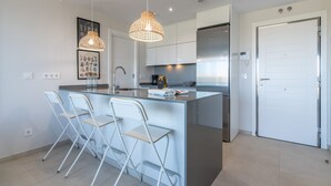 Et moderne køkken med en central ø, barstole, hængende lamper og et rustfrit stål køleskab.