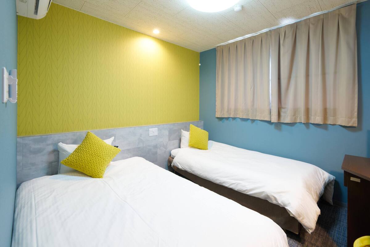 A hotel room with two beds, a ceiling fan, a window with curtains, and a wall-mounted air conditioner.