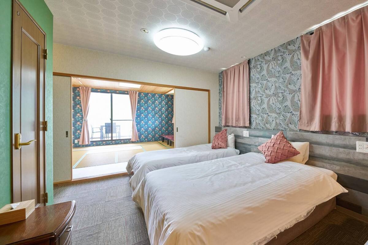 A hotel room with two beds, a wooden desk, a large window with curtains, and a patterned wall.