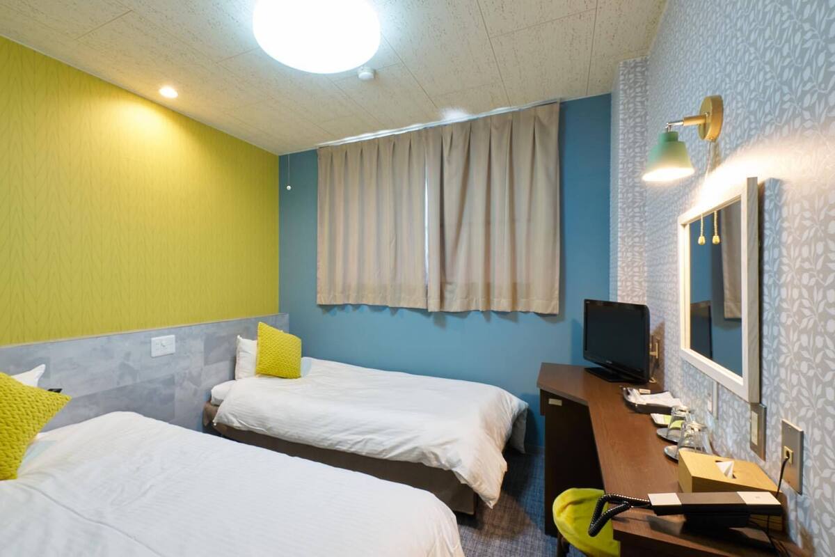 A hotel room with two beds, a desk with a computer, a TV, and a window with curtains.