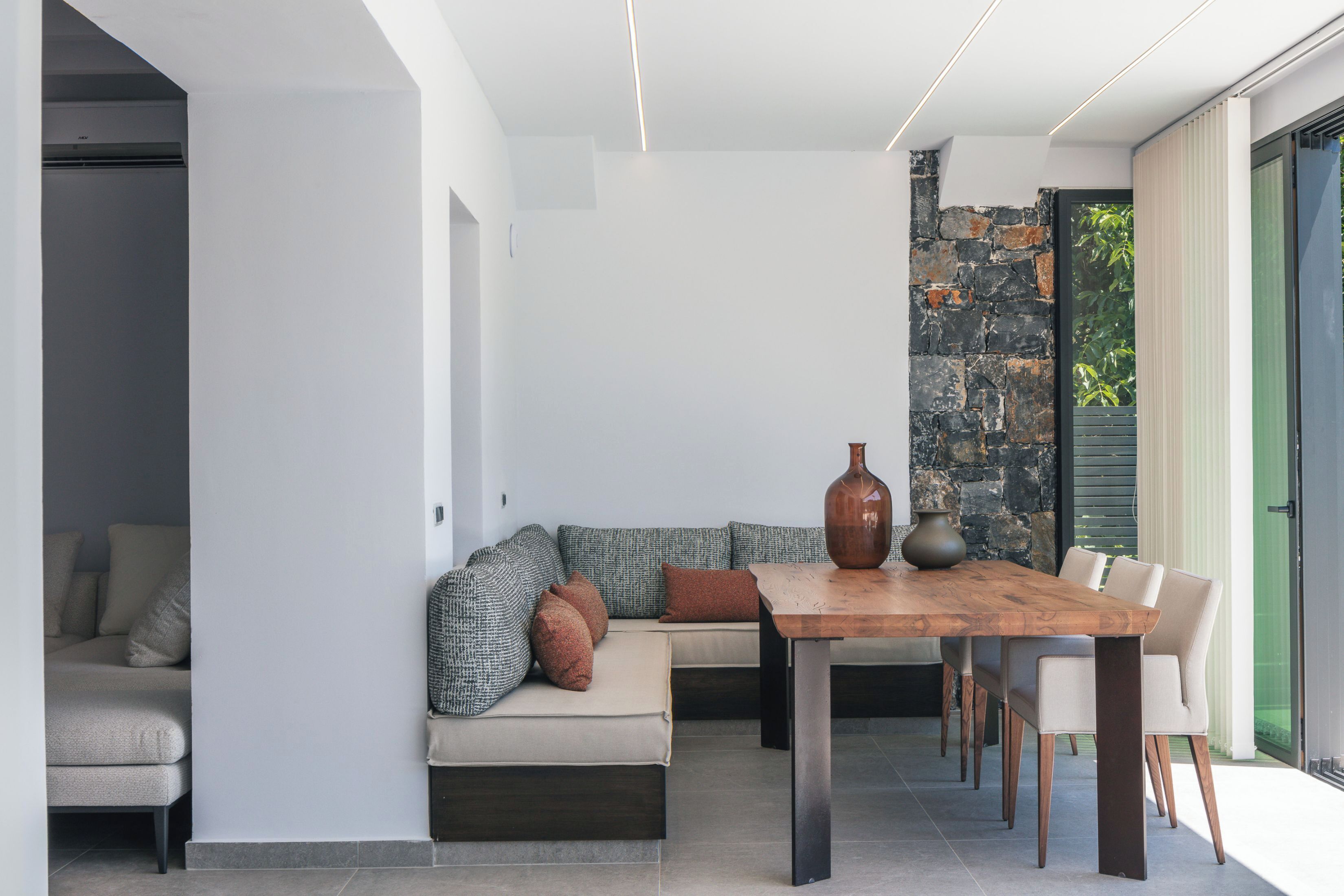 Un'area pranzo moderna con tavolo e sedie in legno, una parete decorata con pietra e vista sul verde attraverso una porta in vetro.