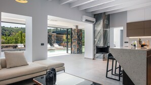 A modern living room with a sofa, a coffee table, and a fireplace. There is a kitchen area with a marble countertop and a bar. The room has large windows offering a view of greenery and a pool. - Casa del Balo - Harmony in Nature by etouri (Chania)
