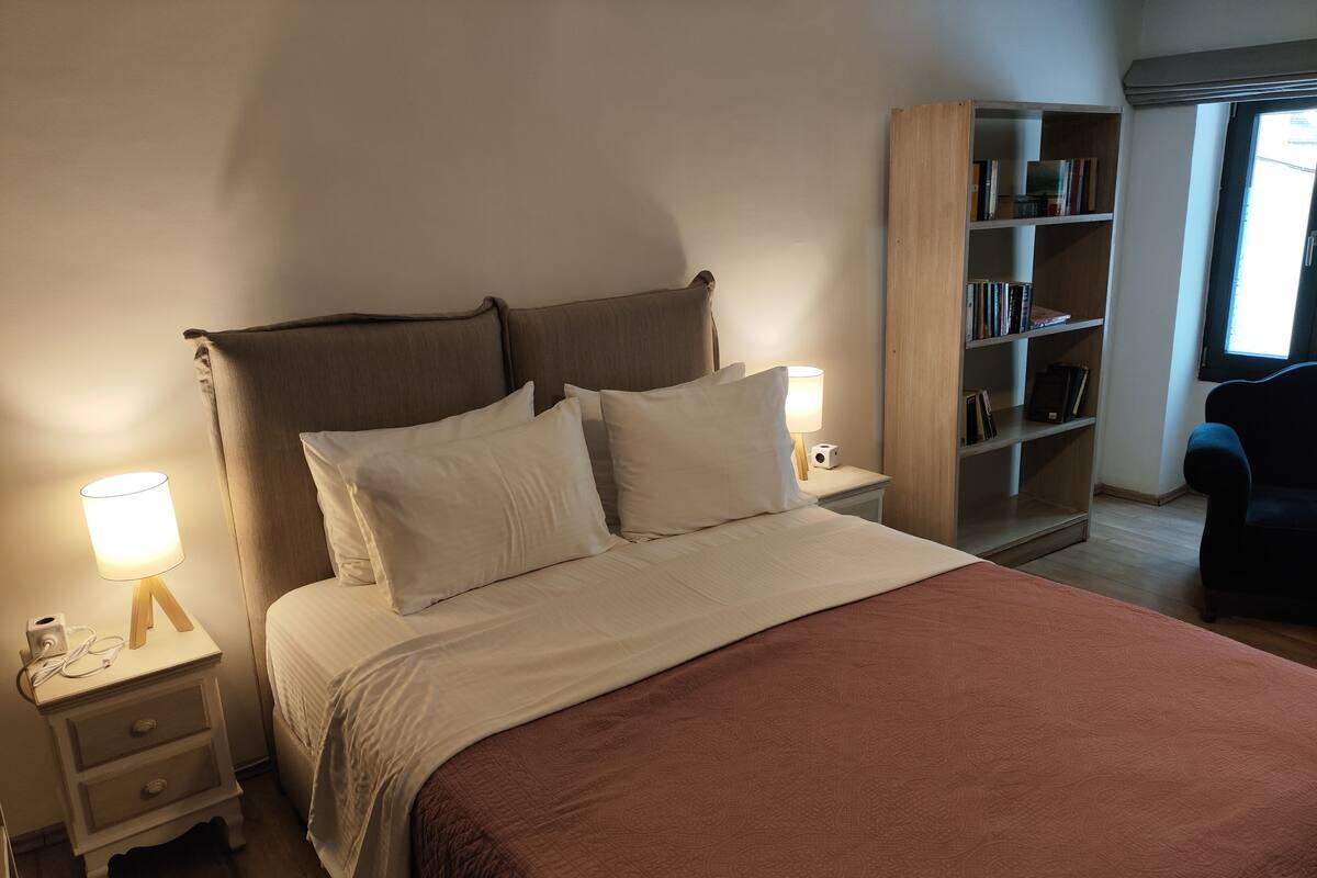 A neatly made bed with a brown blanket, two bedside lamps, a bookshelf, and a chair.