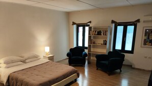 Un dormitorio con cama, mesita de noche, lámpara, dos sillones, una estantería y una ventana con cortinas.