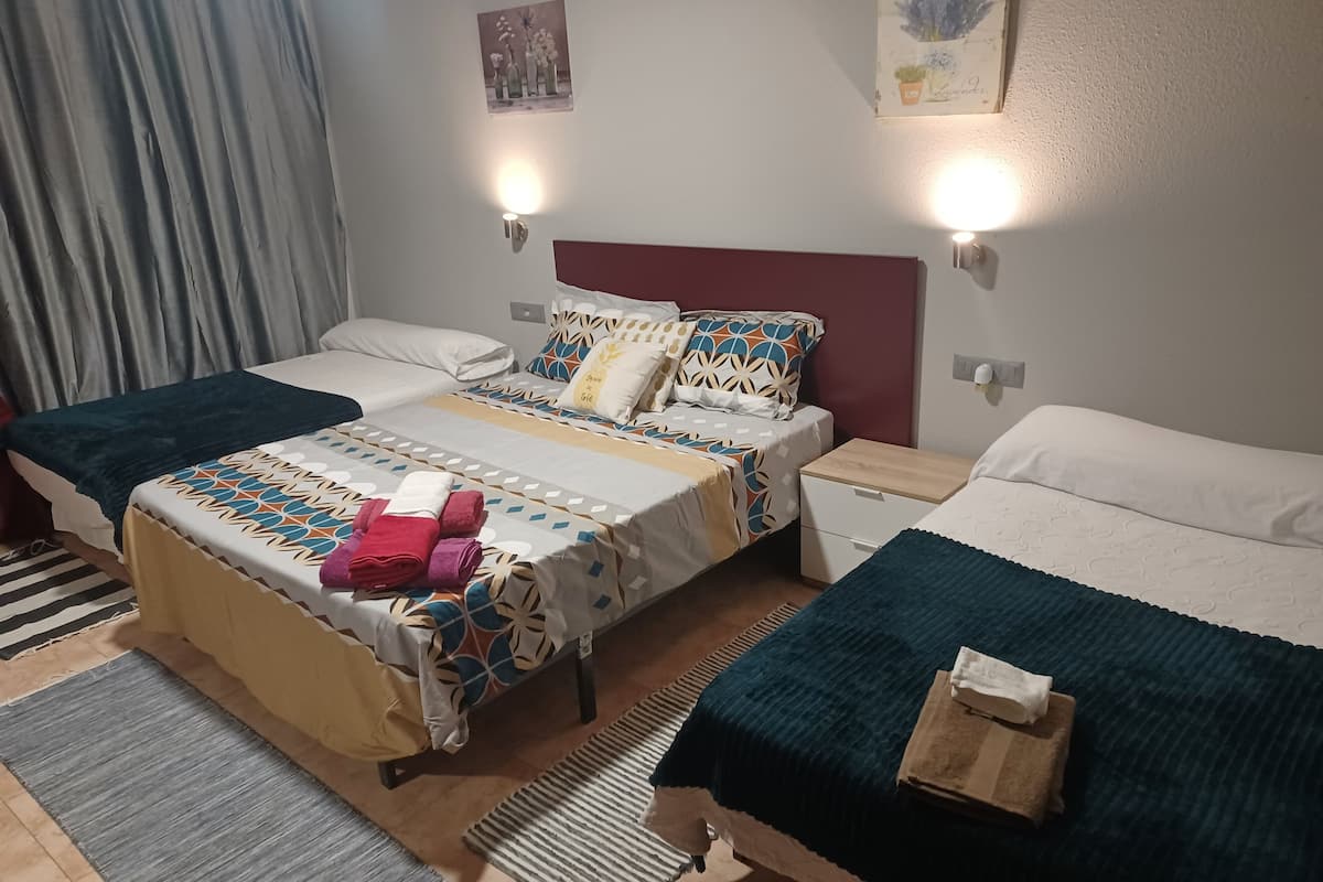 A hotel room with two beds, a large bed with a patterned bedspread and pillows, and a smaller bed with a plain bedspread. There are two wall-mounted lights, a window with curtains, and a small table with a towel.