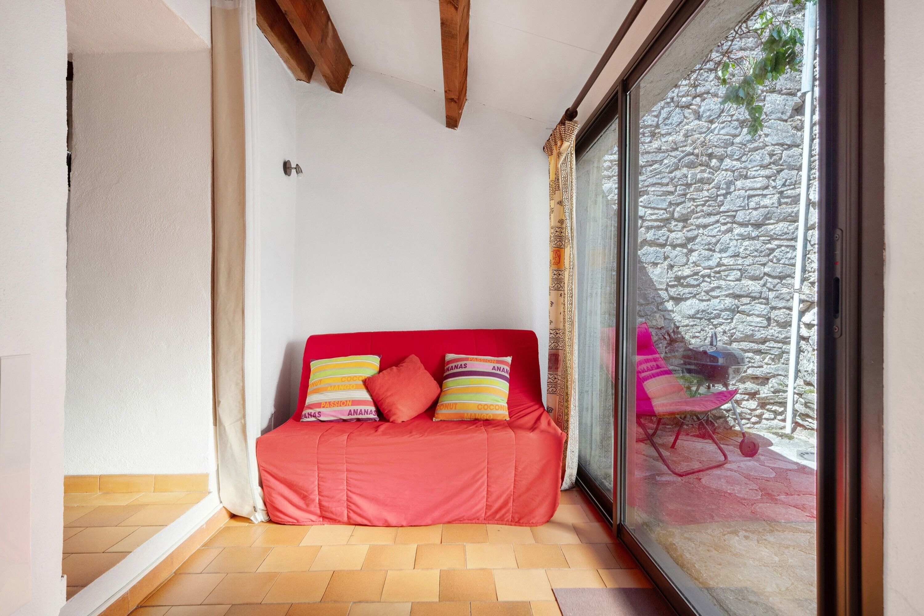 Un canapé rouge avec des coussins à motifs, une porte en verre donnant sur un mur en pierre et un sol carrelé.