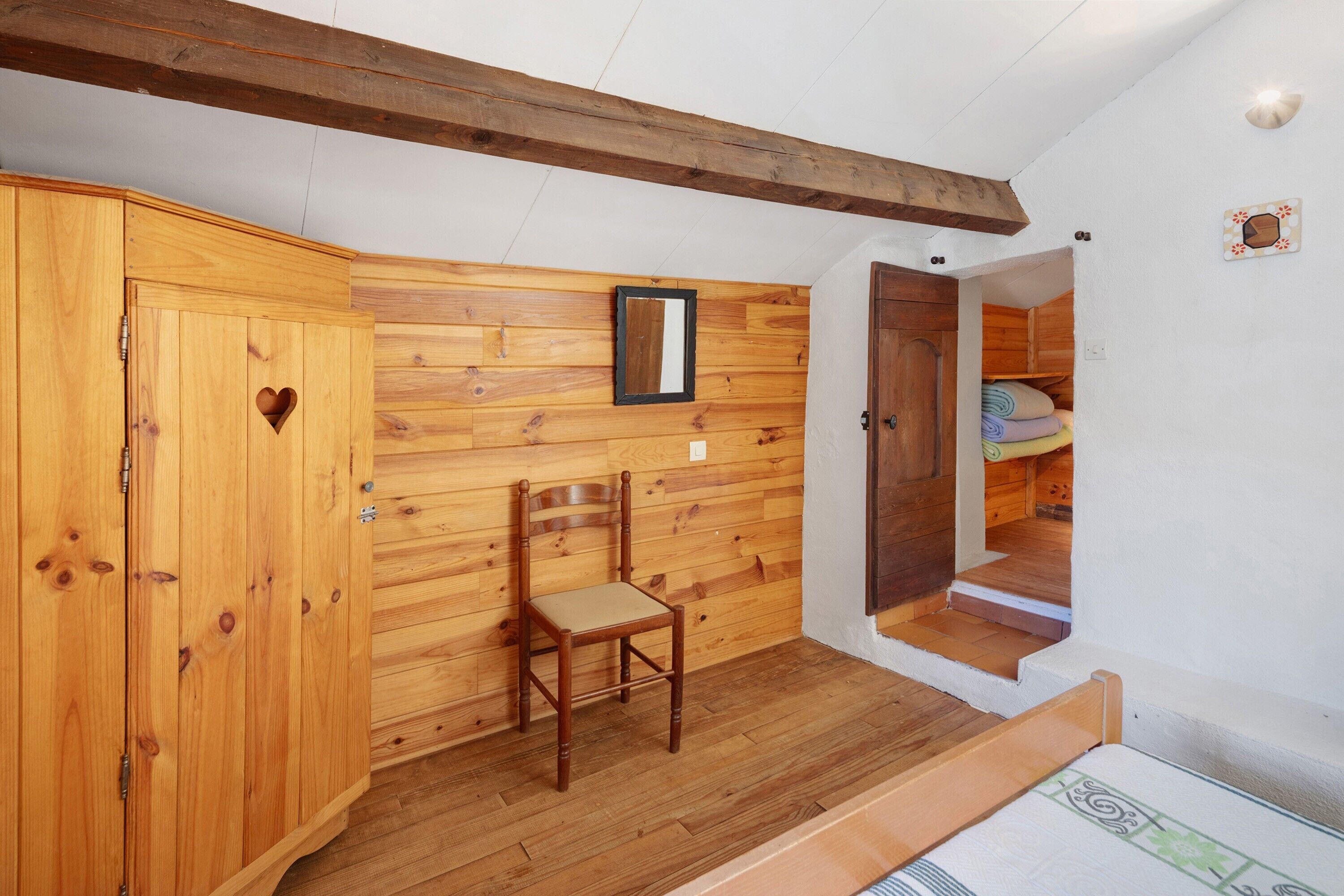 Um quarto com painéis de madeira, um guarda-roupa de madeira, uma cadeira, um espelho e uma escada que leva a outro cômodo.