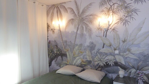 Un dormitorio con un mural de follaje tropical, una cama con ropa de cama verde y una mesita de noche con lámpara.