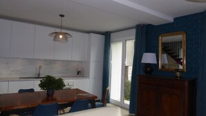 Un comedor con una mesa de madera, sillas, una planta en maceta, una lámpara colgante, un mueble con espejo y vistas a través de una puerta de cristal.