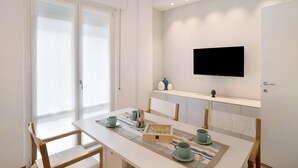 A modern room with a dining table, chairs, a flat-screen TV, and a window with blinds. - Apartment 'Casa Federica' with Balcony and Wi-Fi (Alassio)