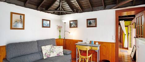 Una habitación con un sofá gris, un escritorio de madera y una silla. Hay cuadros en la pared, una lámpara y un cubo de basura.