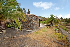 Un bâtiment en pierre avec un toit en tuiles, entouré de palmiers et d’une zone herbeuse aride.