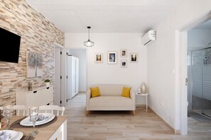Una sala de estar moderna con un sofá, un comedor con una mesa lista para dos personas, y una zona de cocina con una pared de ladrillo y un televisor montado.