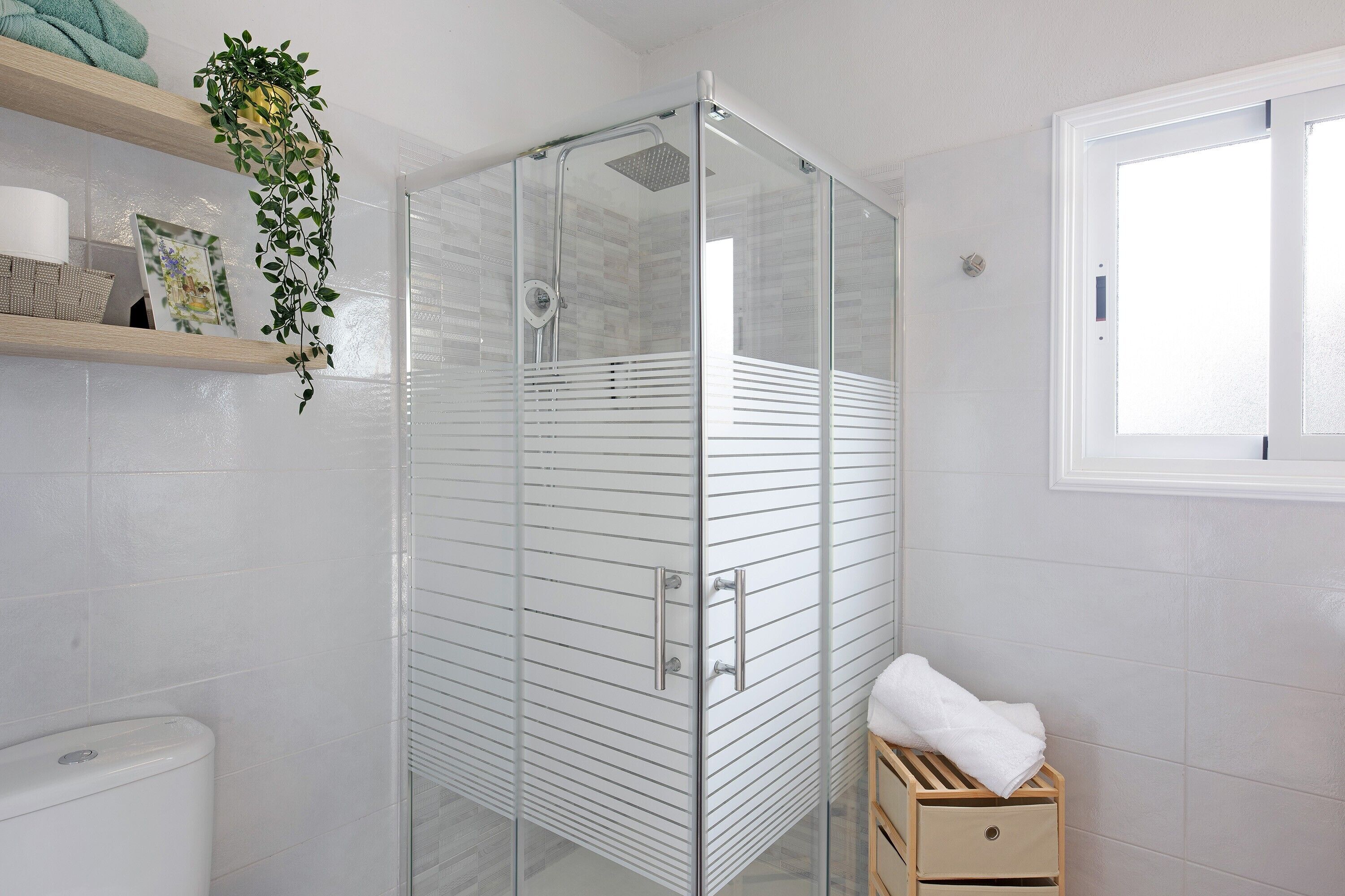 Eine gläserne Dusche mit weiß gefliester Wand, ein Holzregal mit Handtüchern und ein Fenster.