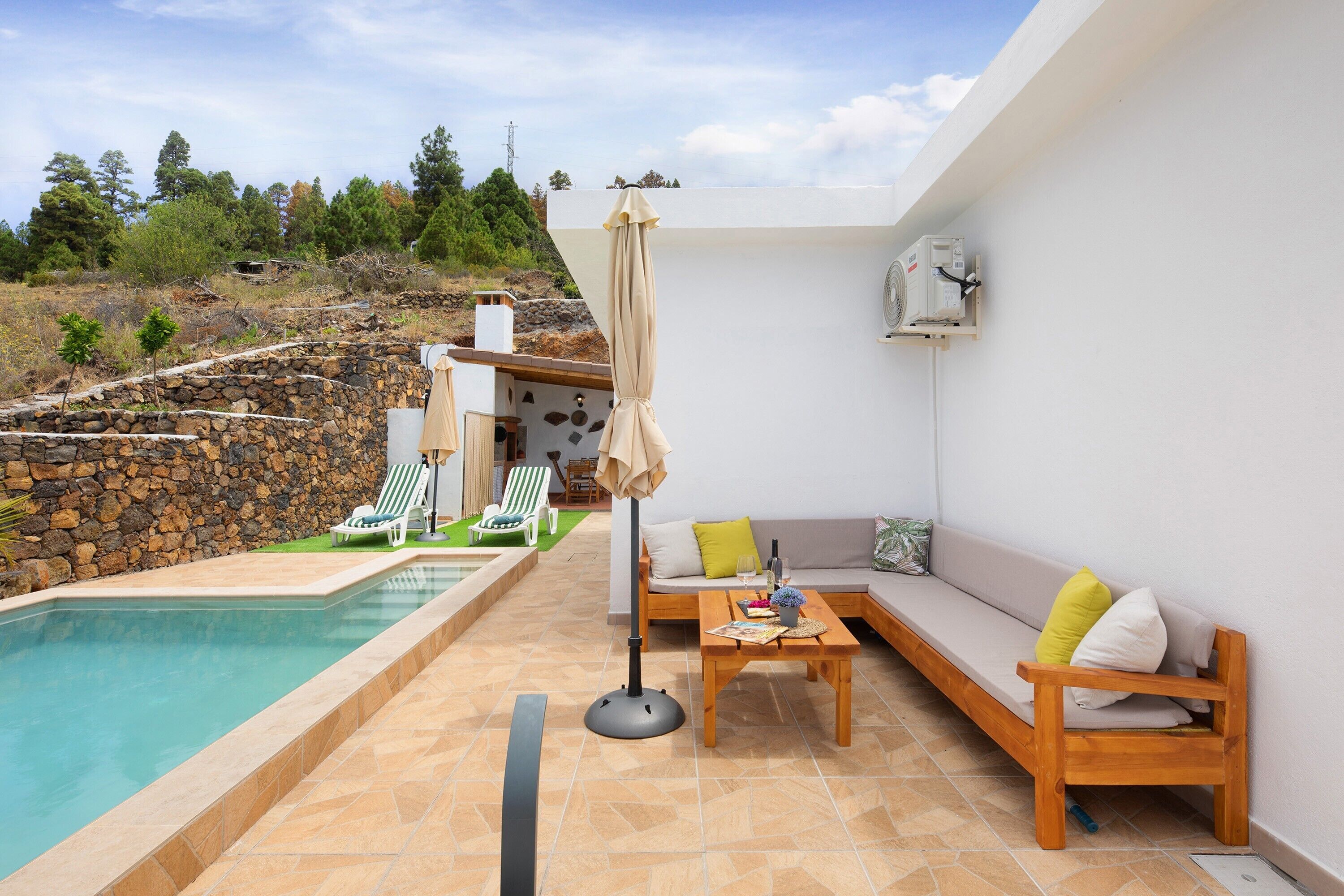 Un espace aménagé au bord de la piscine, avec un mur en pierre, des chaises longues, un canapé et une table sur laquelle se trouvent une bouteille et des verres.