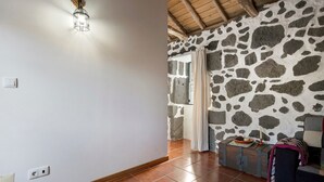 Una habitación con pared de piedra, techo de madera, una lámpara, una ventana con cortinas y una pequeña mesa con una vela.