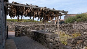 Un muro de piedra, una estructura de madera con hojas secas colgantes, y un edificio de piedra al fondo.