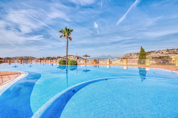 A large outdoor swimming pool with clear blue water, surrounded by a fence, palm trees, and a hilly landscape in the background.