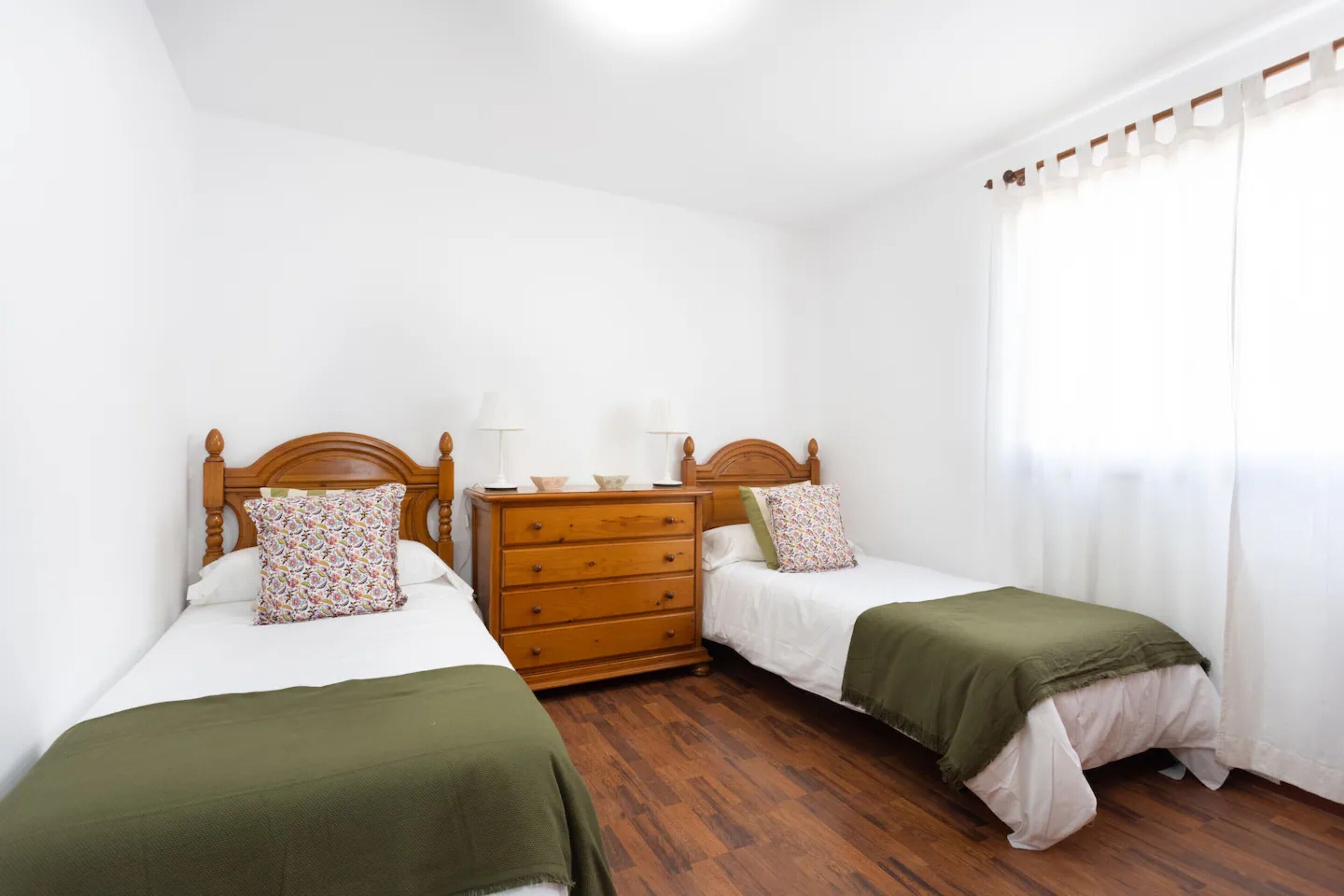 A bedroom with two single beds, a wooden dresser, and a window with curtains.