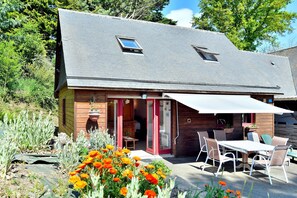 Una casa de madera con techo inclinado, un patio con mobiliario exterior y un jardín con flores de naranja.