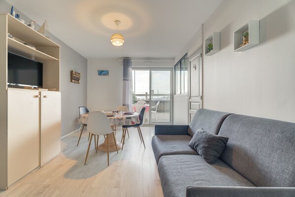 Uma sala moderna com um sofá cinzento, uma mesa de jantar com cadeiras, uma TV sobre um suporte e uma varanda com vista.