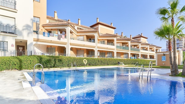 A swimming pool with a tiled edge, surrounded by a hedge, and adjacent to a multi-story building with balconies. - Apartment 'Las Palmeras A92' with Sea Views, Shared Pool and Wi-Fi (MÁLAGA)