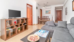 A living room with a flat-screen TV, wooden shelving, a grey sofa, and a dining area with a table and chairs. - Apartment 'Las Palmeras A92' with Sea Views, Shared Pool and Wi-Fi (MÁLAGA)