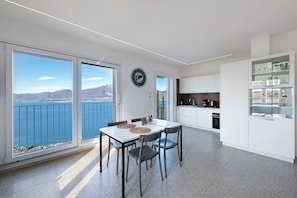 Une cuisine moderne avec des meubles blancs, un coin repas comprenant une table et des chaises, et de grandes fenêtres offrant une vue sur la mer et les montagnes.